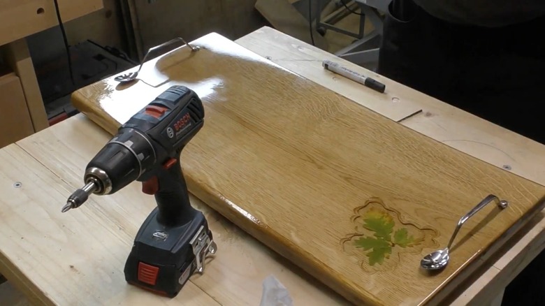 A wooden tray with bent metal spoons for handles sits on a workbench with an electric drill.