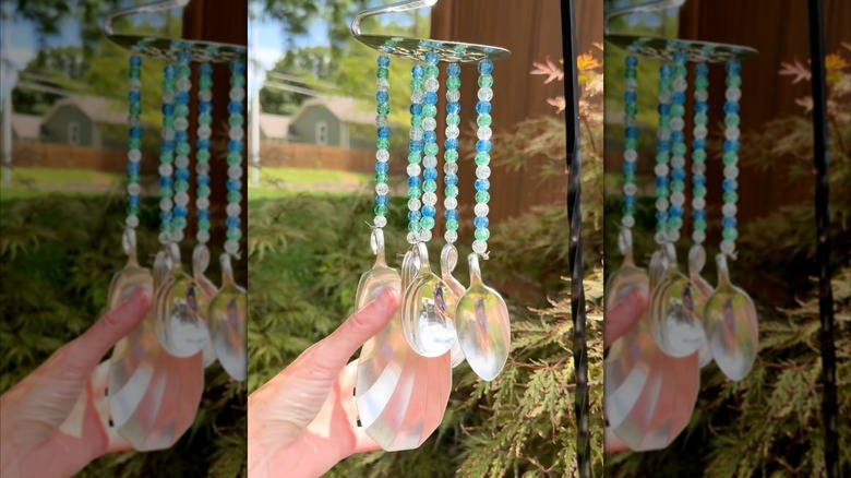 A person holds a wind chime made from metal spoons and beads in their hand while it hangs outside.
