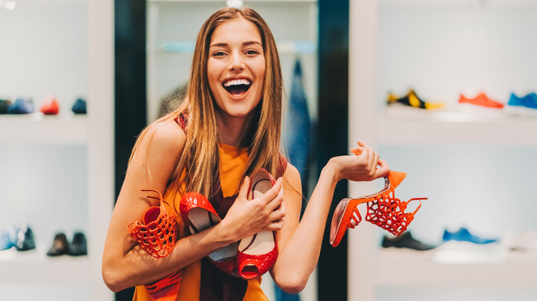 A woman holding multiple pairs of shoes and smiling at the camera. She is wearing an orange and black top and the shoes are orange and red.