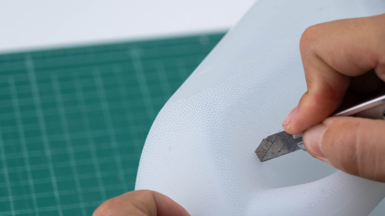A person uses a box cutter to cut into the side of a heavy plastic water jug.