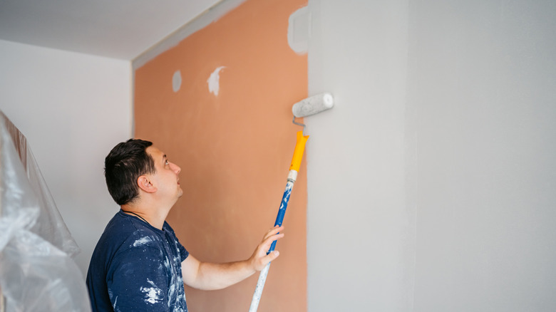 A man uses a roller to paint a wall white in his home.
