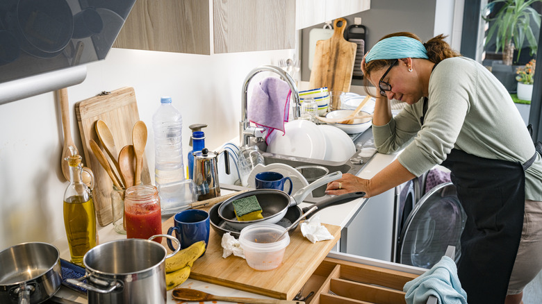 A frustrated woman stands in a kitchen full of dirty dishes spread across the countertop and sink