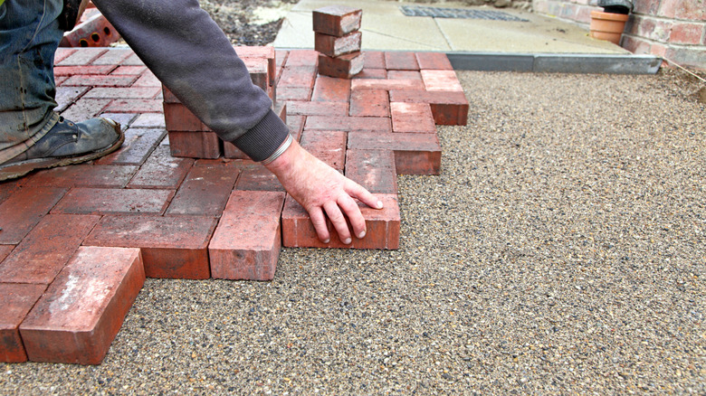 A hand lays red brick pavers on the ground for a driveway
