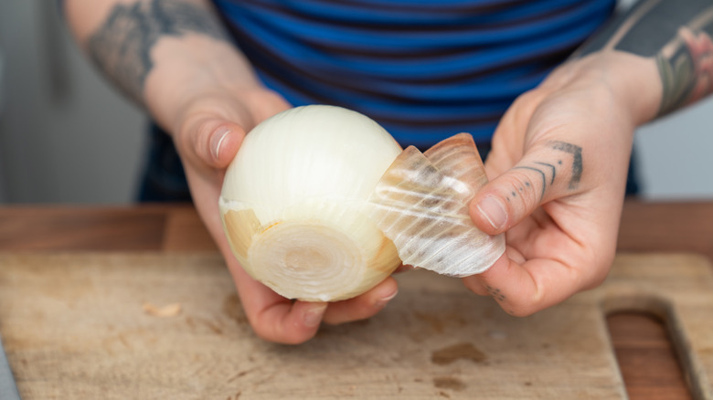A person peels a cut yellow onion over a wooden cutting board.