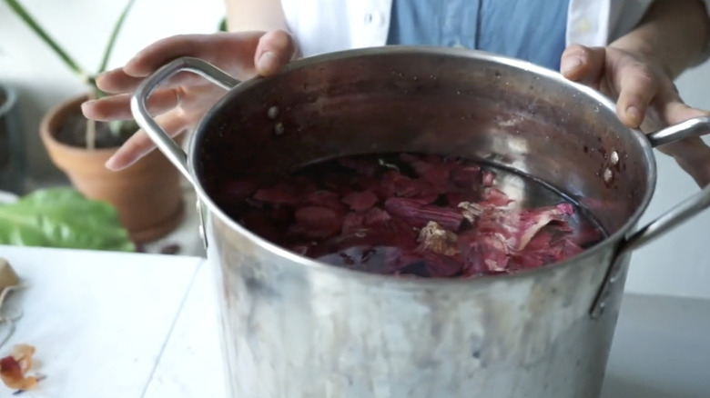 A person makes dye in a metal pot with red onion skins.