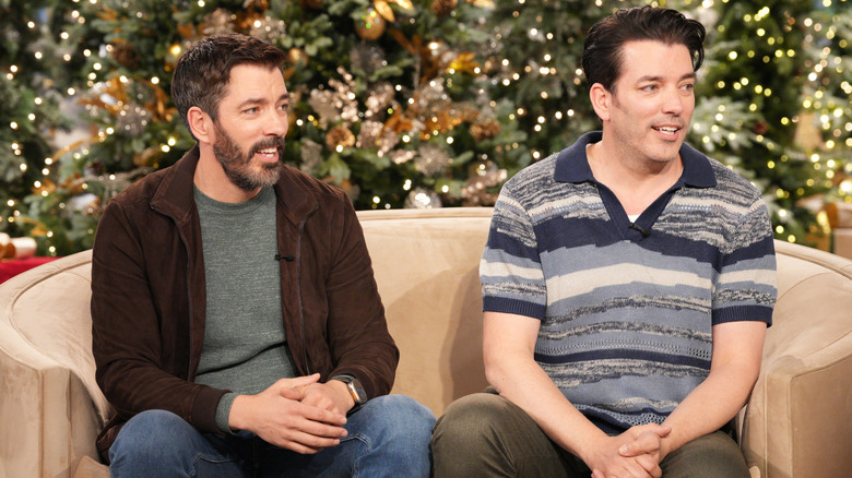 Drew and Jonathan Scott sitting on a tan couch in a studio with Christmas decorations.