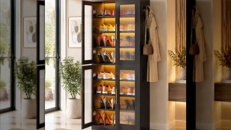 A tall narrow cabinet, with yellow backlit shelves holding rows of shoes, is set against a wall in a bedroom.