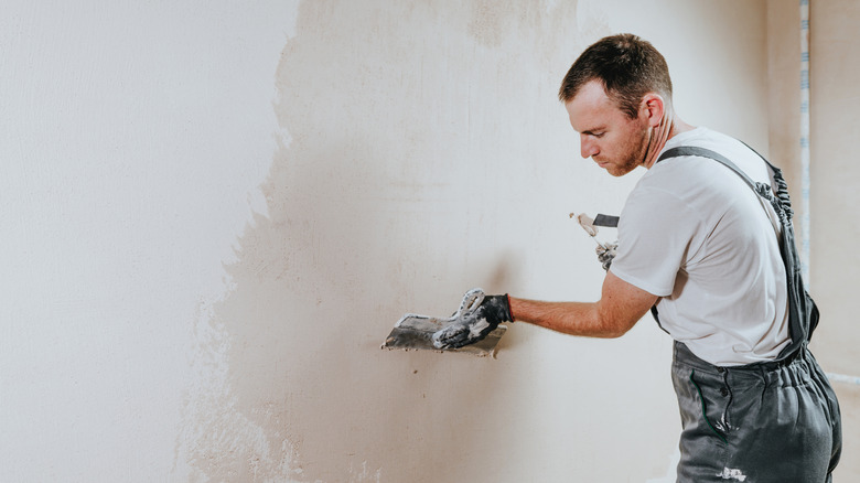 Male builder in work overalls plastering a wall using a construction trowel.