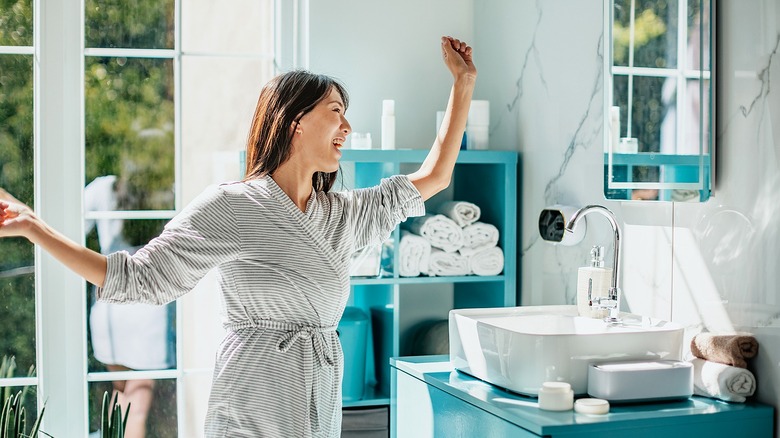 Person smiling and in mid-dance in a white marble bathroom with aqua and white fixtures