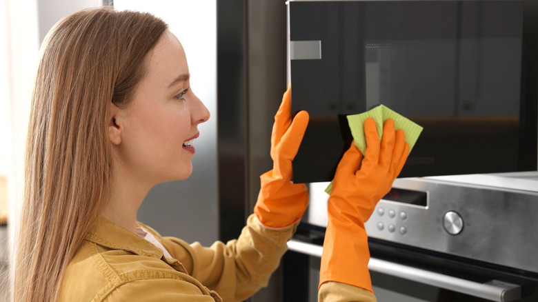 A person wiping down a microwave door while wearing orange rubber gloves