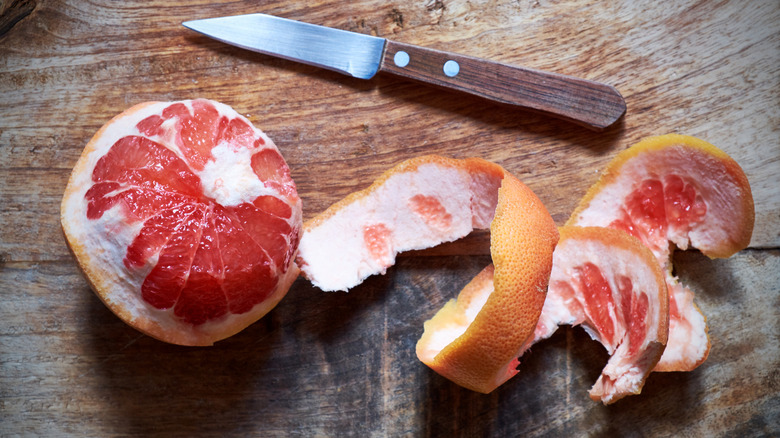 Fresh whole grapefruit partially peeled next to a paring knife on top of a wooden cutting board
