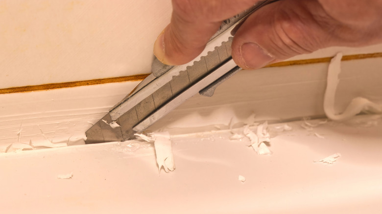 Hand using a utility knife to remove old caulk from tub in bathroom.