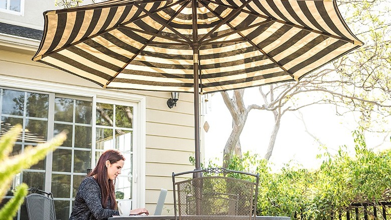 A person works on their laptop outside while being shaded with a striped patio umbrella