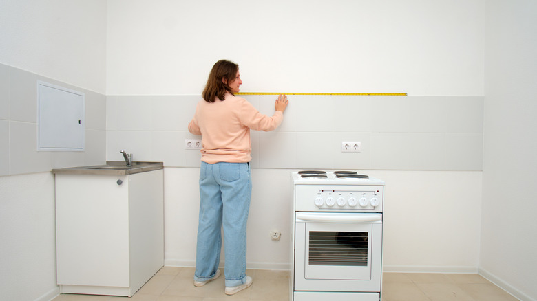 A woman measures the space above a freestanding stove in a kitchen with a yellow measuring tape.