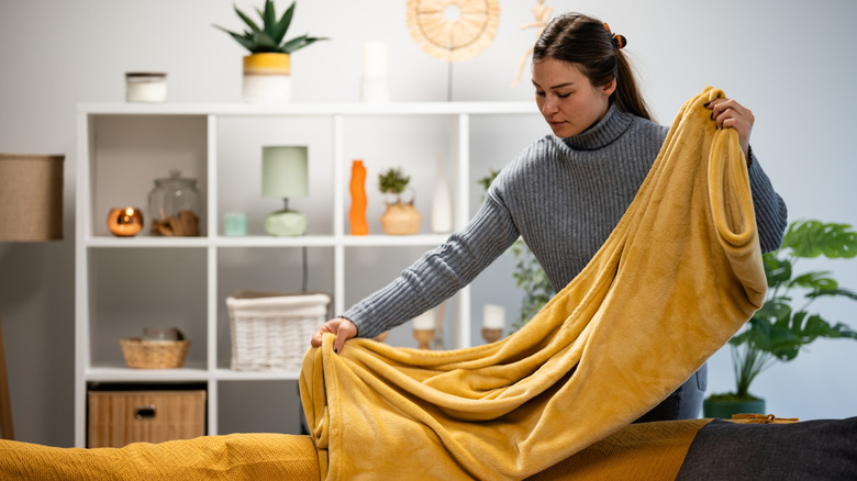 A woman places a folded yellow blanket over the back of a sofa in a living room.