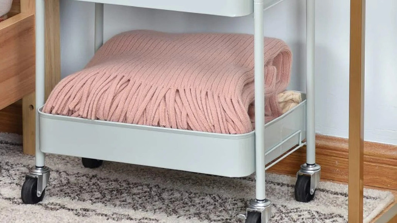 A white metal utility cart on wheels with a pink woollen blanket folded up in the bottom shelf.