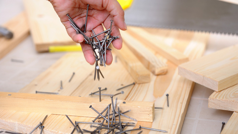 A hand drops a bundle of nails onto a workbench covered in pieces of wood and carpentry tools.