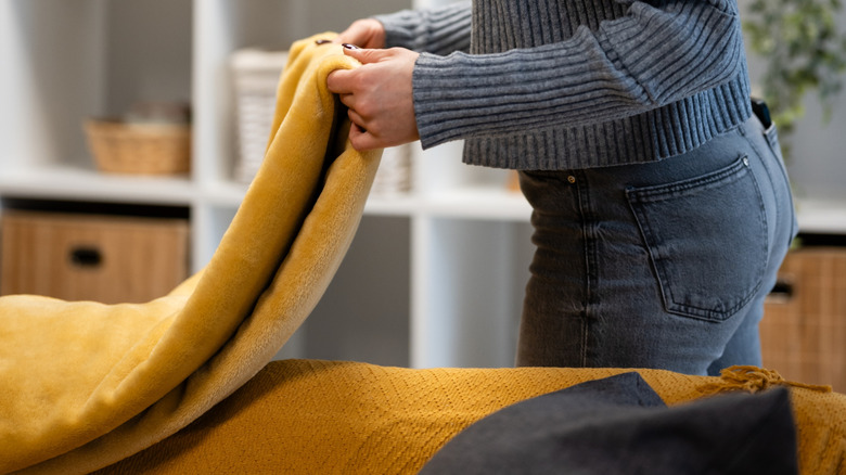 A person lays a neatly folded soft yellow blanket over the back of a sofa.