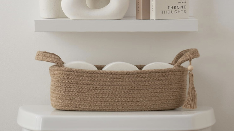 Jute basket used as storage for toilet paper, placed on top of the toilet tank and below a floating shelf adorned with decor and books