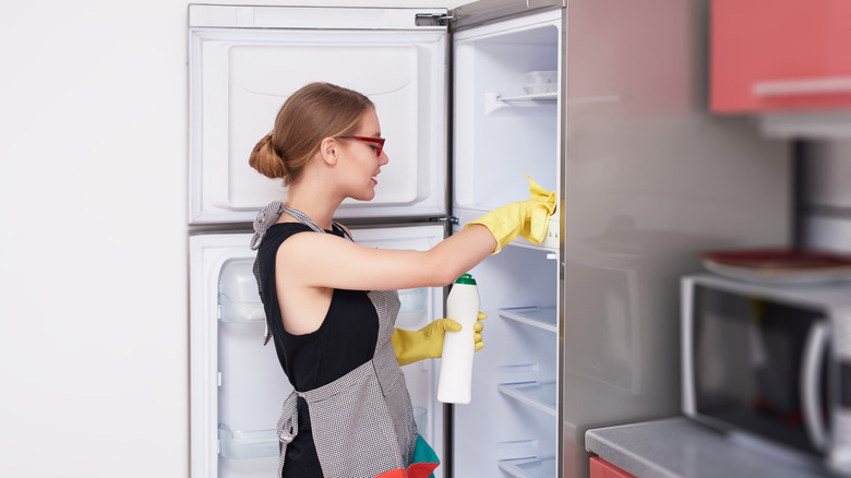 A woman cleaning out her empty fridge and freezer wearing an apron and yellow gloves