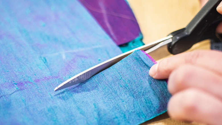 A person holding black scissors cutting into a blue and purple piece of fabric