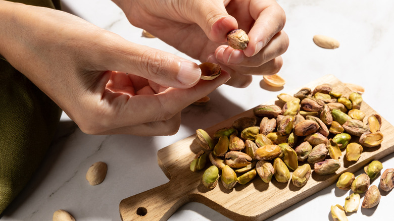 A person peeling off shells from pistachios on a small wooden cutting board