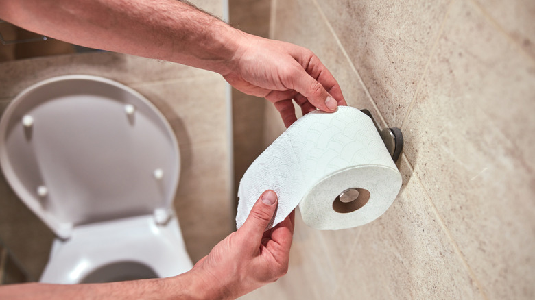 Two hands remove toilet paper from a roll that's hanging on a wall-mounted holder.