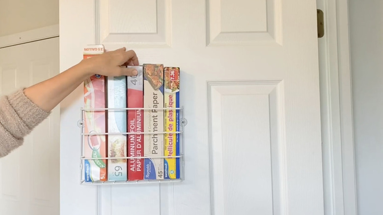 A person reaches for a box of aluminum foil stored in an Evelots organizer mounted on a door in the kitchen.