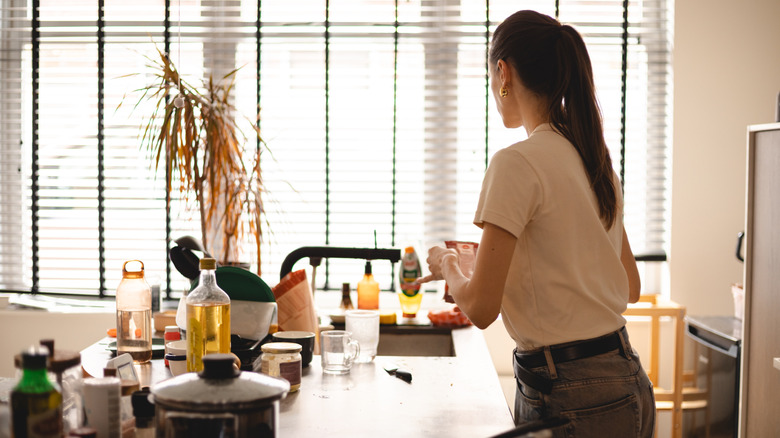 A woman examines a food packet while standing in a kitchen with a cluttered countertop.