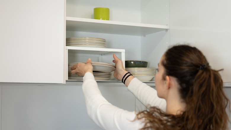 A woman placing white dishes into an open white dish cabinet