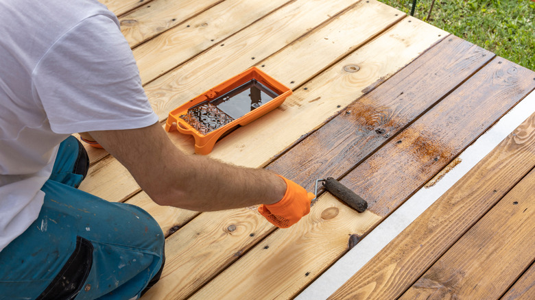 A person applies a dark stain to a newly completed wooden deck with a small paint roller.