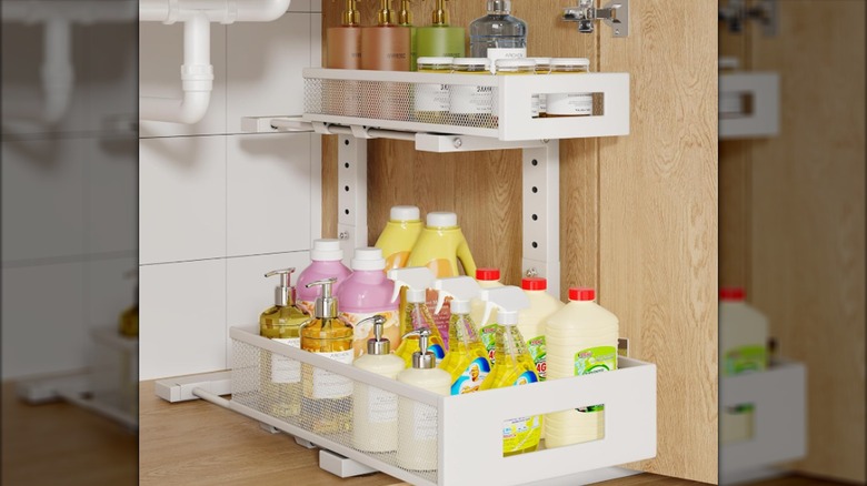 A white two-tier pull-out shelf unit holds essentials in a cabinet under a sink.