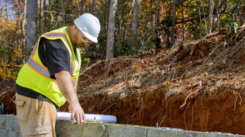 Construction worker measuring a retaining wall being built beside a dirt hill.