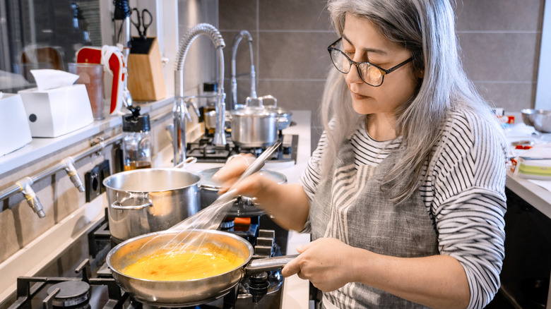 Older woman whisks eggs in a stainless steel pan in a restaurant kitchen.