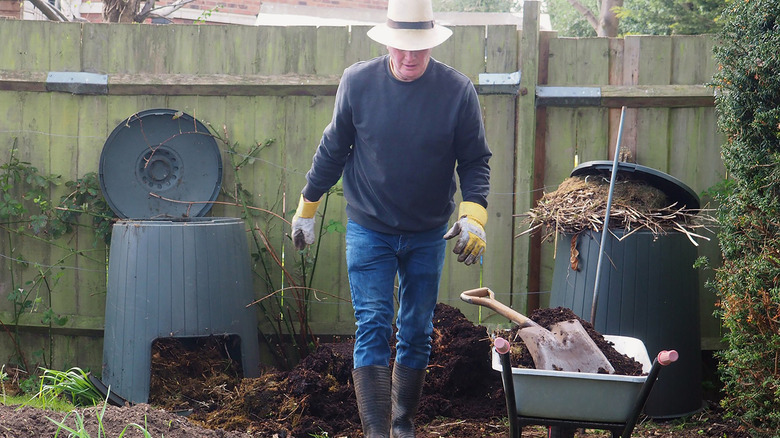 A man wearing gardening gloves walks away from two large black plastic compost bins in his backyard.