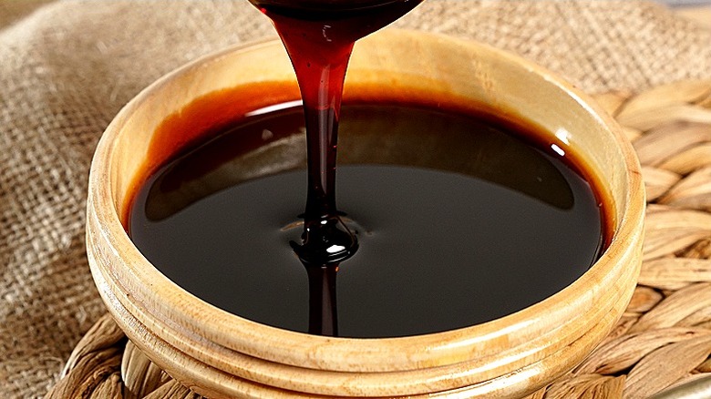 A dark syrupy liquid is dripping off a wooden spoon into a light wooden bowl that sits on a woven straw mat