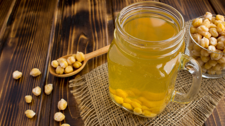A glass jar with chickpeas and chickpea water in it, next to a cup of chickpeas in the background and scattered on the wooden table