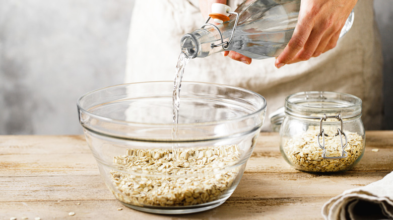Person pouring a bottle of water into a glass bowl filled with uncooked oats.