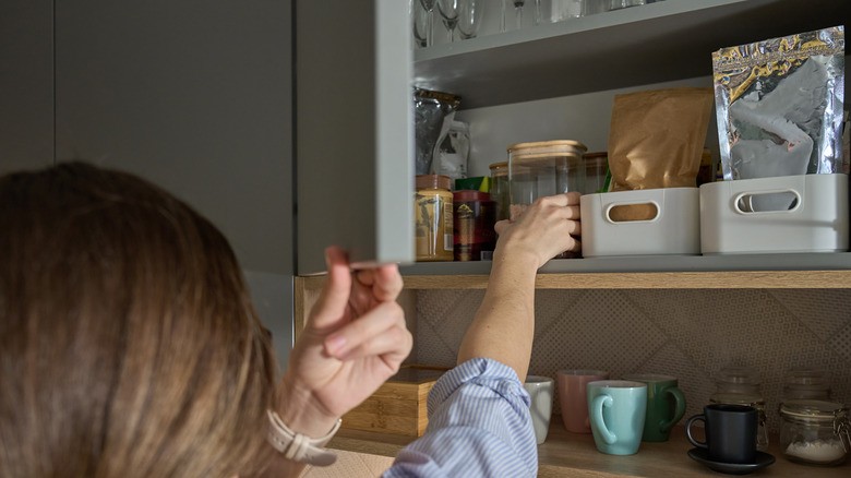 A woman reaching into a kitchen cabinet that contains glass canisters and packaged items.