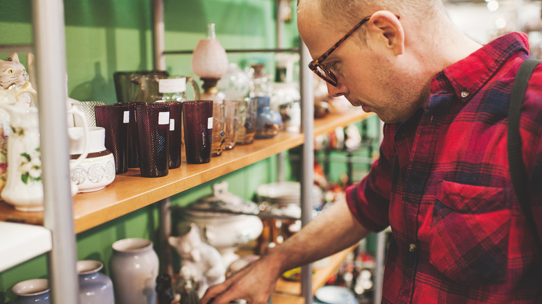 Man in red checkered shirt and tortoiseshell pattern glasses reaches for a ceramic on a thrift store shelf.