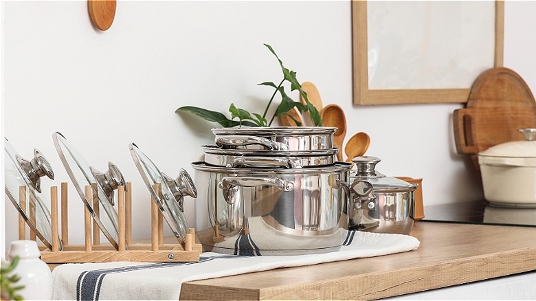 Stainless steel cookware set sits on kitchen countertop with lids stacked in wooden holder