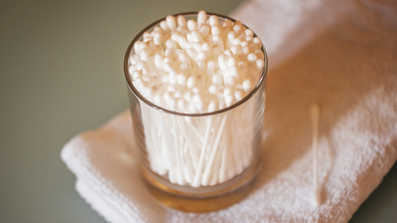 Glass jar containing cotton swabs rests on a white towel, with one cotton swab positioned next to the jar on a flat surface.
