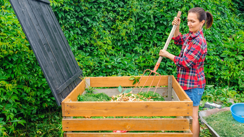 A woman in a red plaid shirt is turning compost ingredients with a pitchfork in a wood-sided compost bin