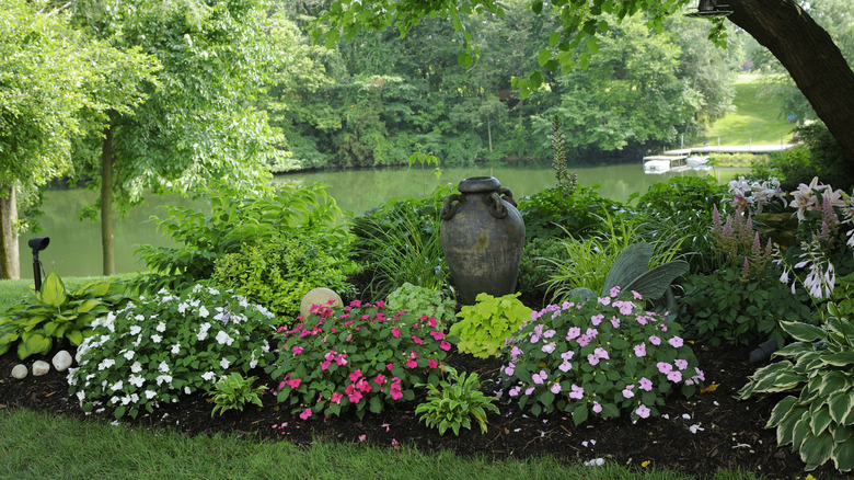 A shaded garden bed planted with flowering plants and shrubs borders a large pond.