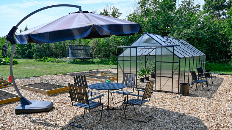 A backyard patio with a wood chip flooring houses a patio table and chairs, a greenhouse, and a large outdoor umbrella.