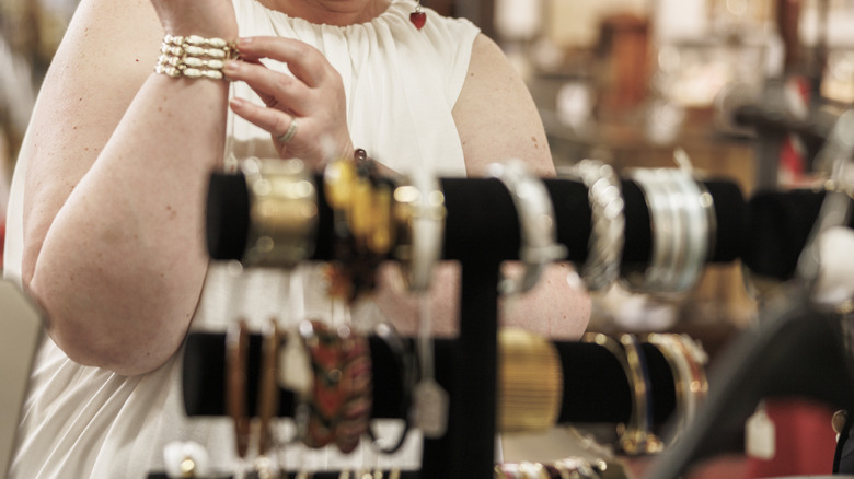 Person standing in a thrift store and trying on jewelry with a jewelry stand blurred in the foreground
