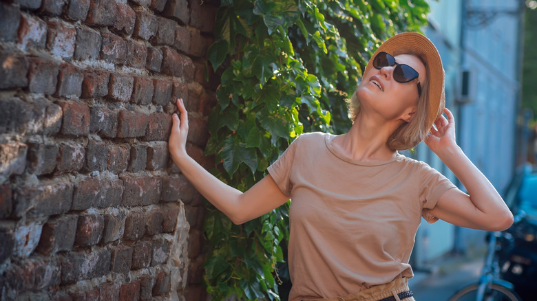 A woman in hat and sunglasses is looking at a blank stretch of brick wall with vines behind her.