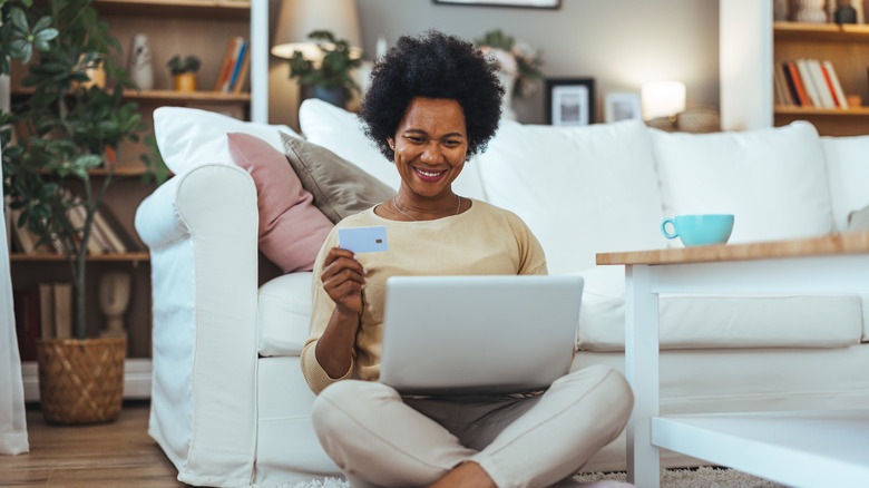 A woman sits near sofa using laptop with a credit card in the other.