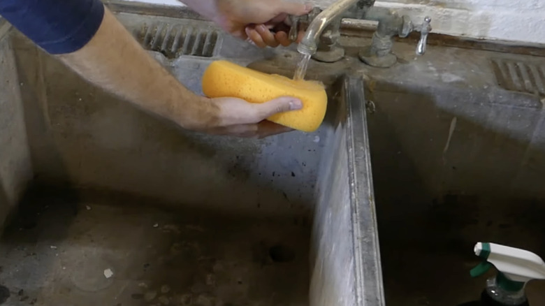 A man cleans a vintage farmhouse-style double concrete sink with a yellow sponge and spray-on cleaner.
