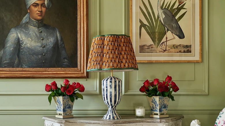 Penny Morrison Blue on Cinnamon Marigold Pleated Silk Scalloped Lampshade with Green Trim on a blue and white striped ceramic base, styled on a side table with rose vases. Portrait and nature paintings on a green wall in the background.